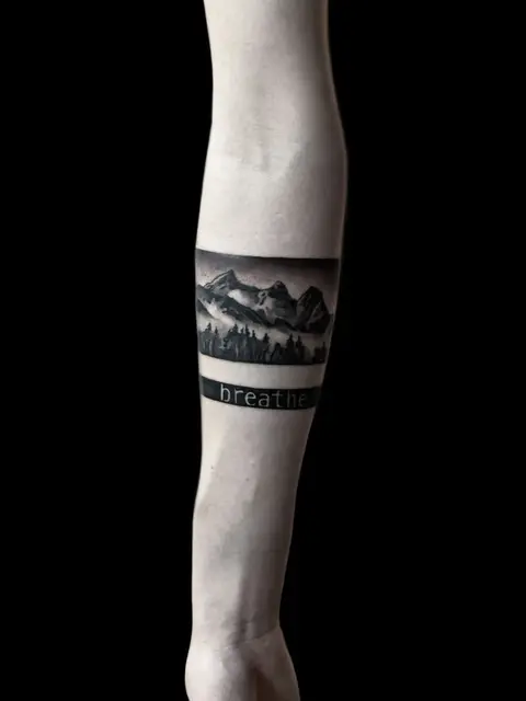 Berglandschaft mit Nebel und Tannen in Black & Grey am Unterarm, darunter Schriftzug «breathe»